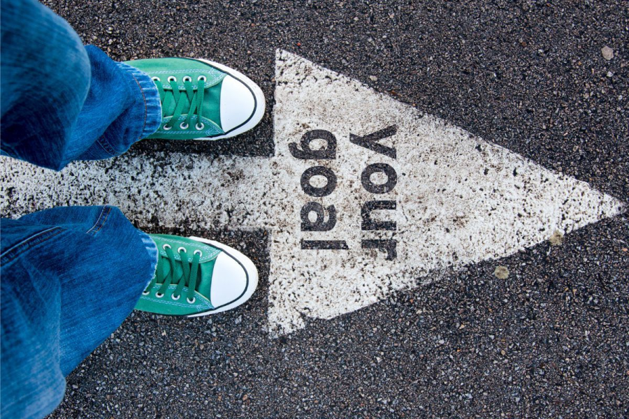 A person's feet standing on a roadway with the text "your goal" in a white arrow on the ground.
