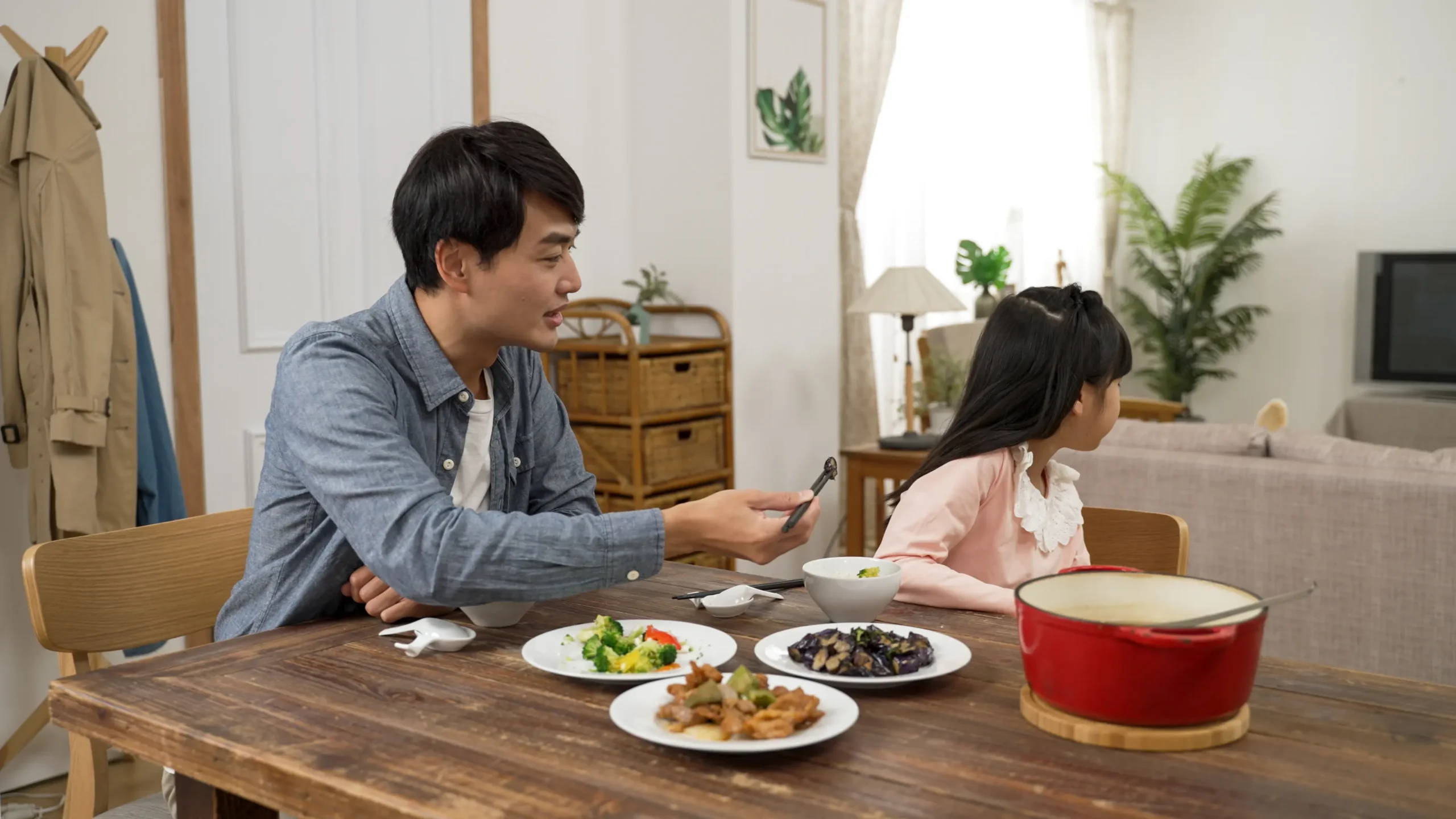 Upset asian daughter turning head and refusing to eat while her father is feeding her vegetable food with chopsticks in the dining room at home
