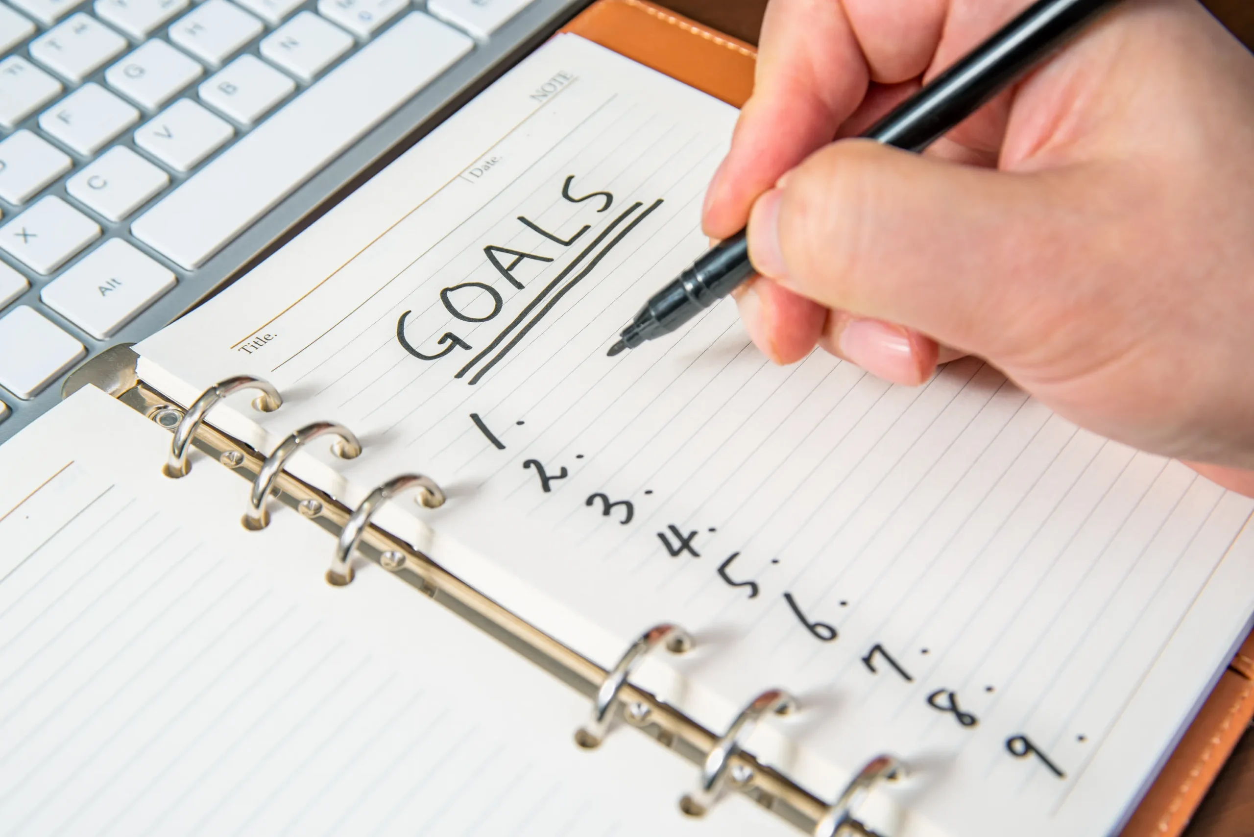 Closeup of a notebook with "Goals" at the top, numbered lines below, and a hand getting ready to start writing a list